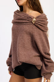 A woman wearing a heathered brown off-the-shoulder knit sweater with a large twisted detail and a gold-tone ring clasp, paired with black shorts.