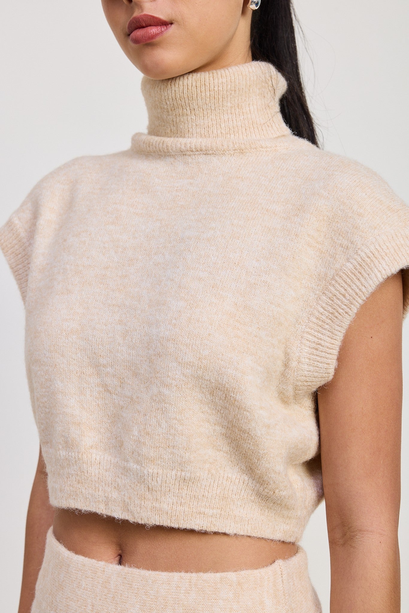 A close-up of a model wearing a soft, fuzzy cropped knit turtleneck sweater in a light cream or oatmeal color.